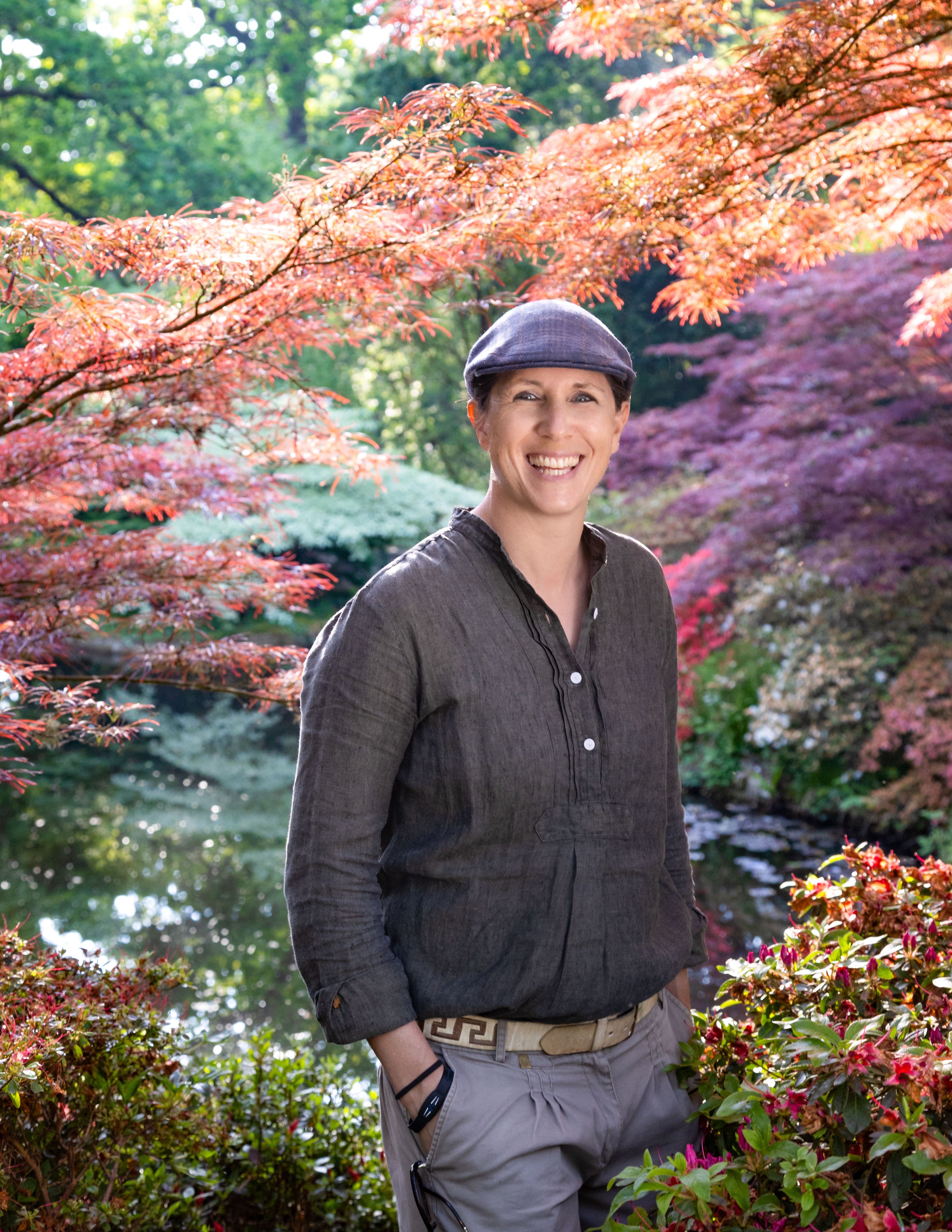 Marie-Louise Agius standing beneath a coral-leaved Japanese maple beside a still garden pool